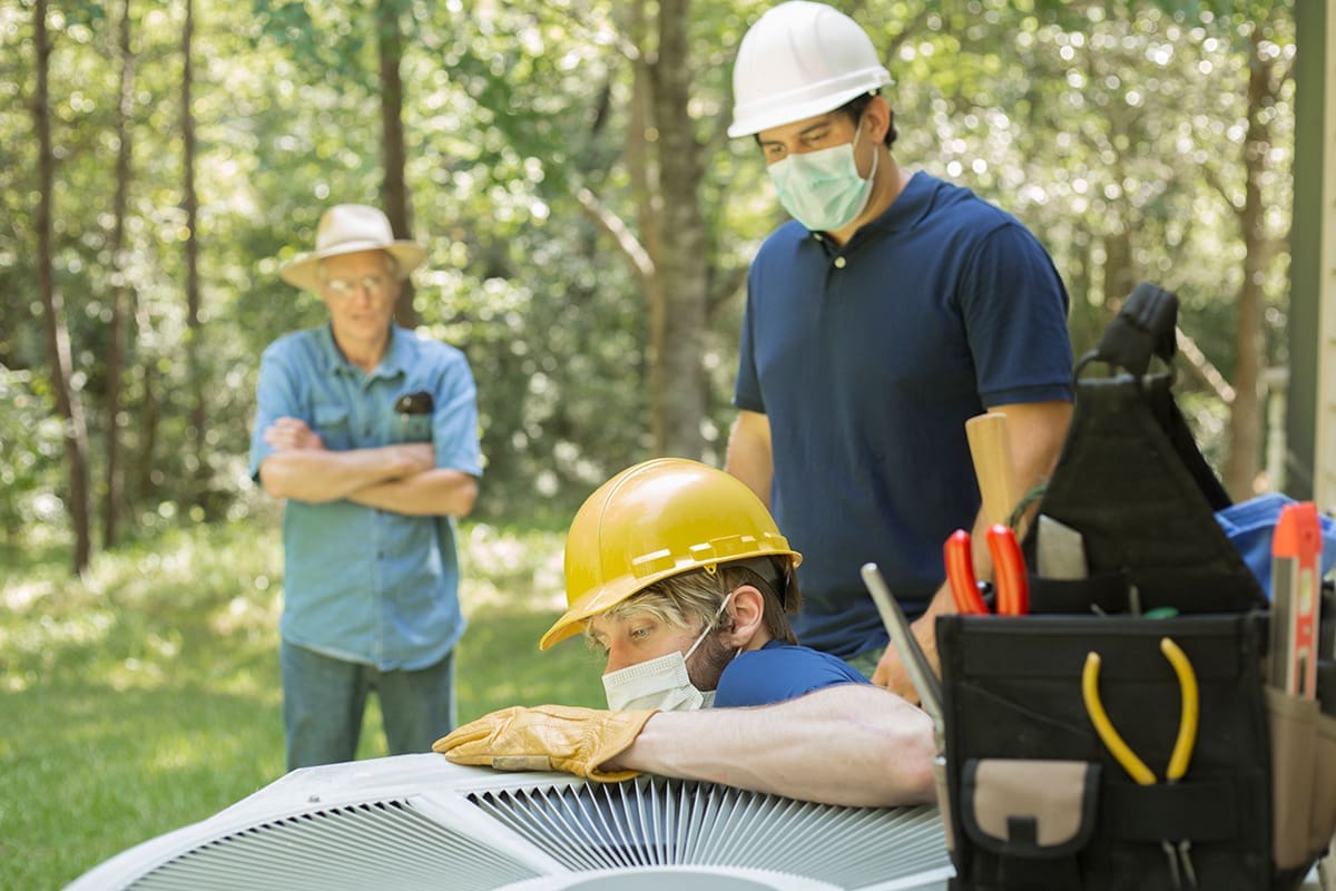 A pair of service men working on an A/C unit with a customer watching in the background